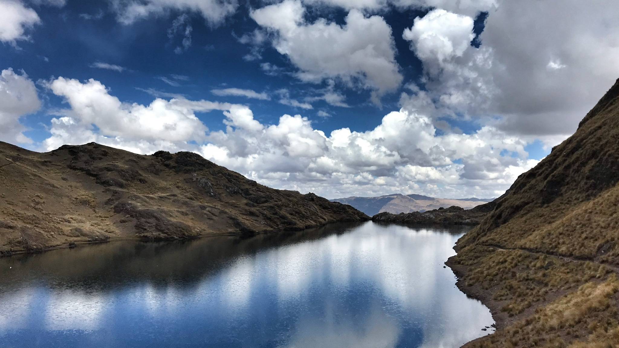A still high mountain lake reflecting a sky of broken white cloud, dry slopes rising on both sides