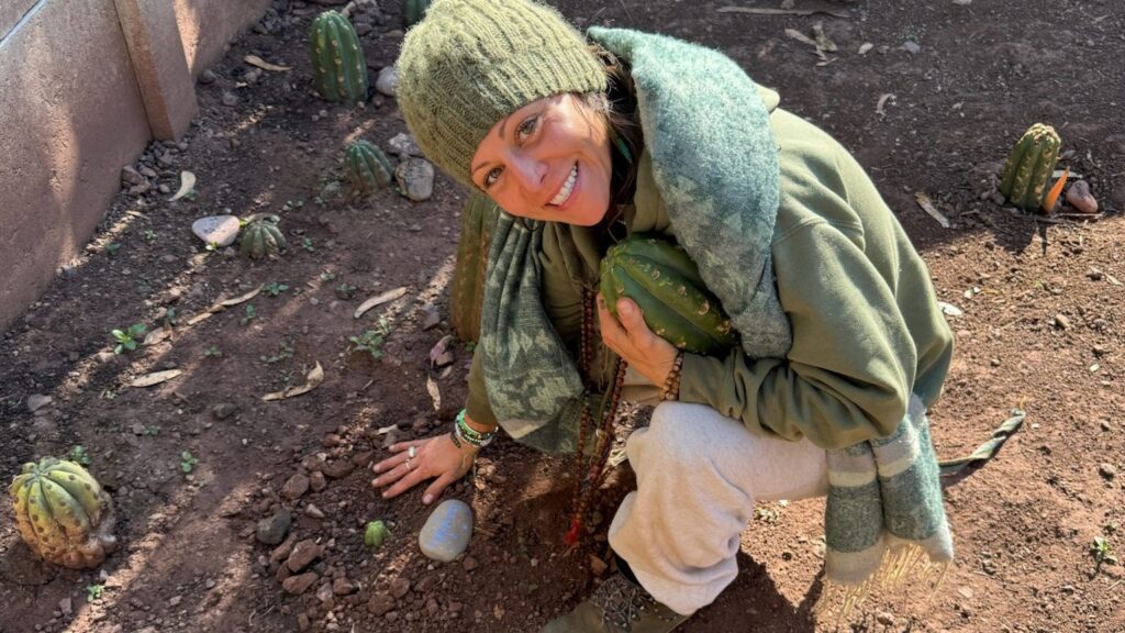 A person kneeling in the garden holding a cut Huachuma segment against their chest, planted cuttings around them