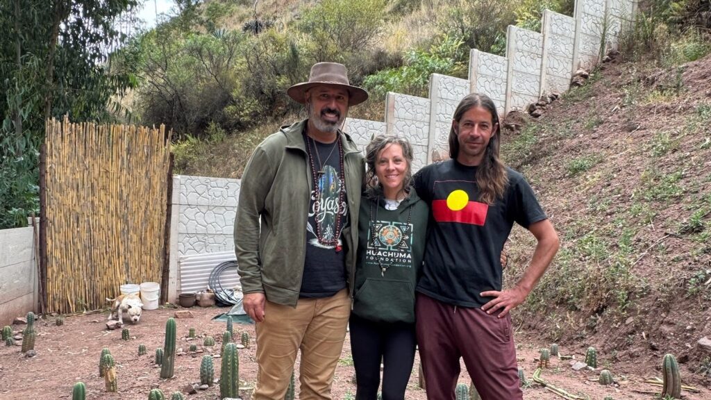 Three people standing together among rows of young Huachuma plants, the garden wall climbing the hill behind