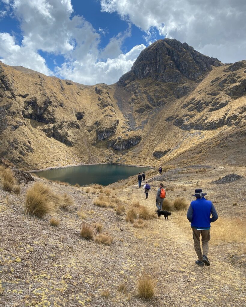A line of walkers descending a tussock slope toward a dark lake in a bowl below a rocky peak, a dog running with them