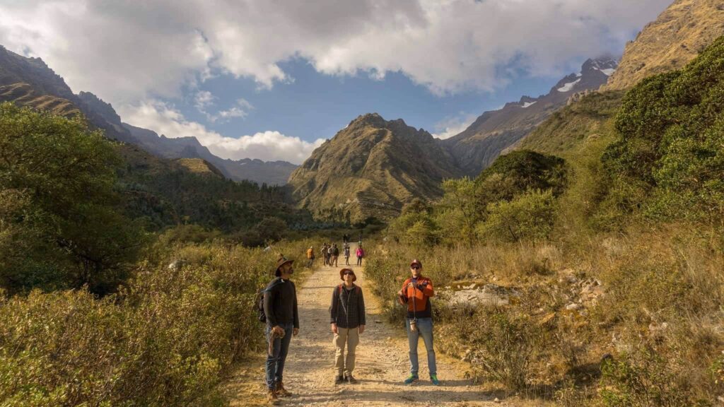 Three people standing on a gravel track in a high mountain valley, more walkers ahead of them and snow on the peaks