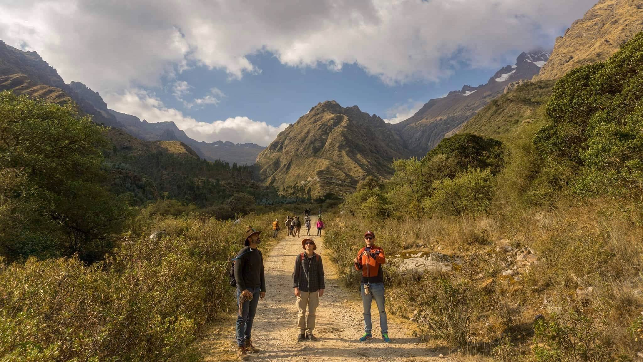 Three people standing on a gravel track in a high mountain valley, more walkers ahead of them and snow on the peaks